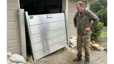Homme debout dans l’eau devant un garage protégé par un batardeau en aluminium pendant une inondation, avec sacs de sable autour de la barrière