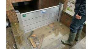 Batardeau en aluminium retenant l’eau boueuse devant un escalier de sous-sol pendant un épisode de pluie, avec une personne en bottes inspectant la barrière anti inondation
