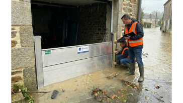 Deux techniciens en gilet orange contrôlent un batardeau aluminium installé devant un garage pendant une inondation de rue