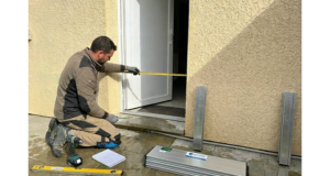 Technicien à genoux prenant la largeur d’une porte au mètre ruban, avec éléments de batardeau aluminium posés au sol pour installer une protection anti inondation