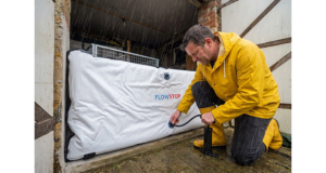 Homme en ciré jaune gonflant un batardeau gonflable Flowstop devant l’entrée de son garage sous la pluie pour se protéger d’une montée des eaux