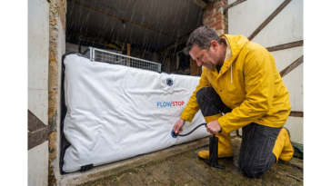 Homme en ciré jaune gonflant un batardeau gonflable Flowstop devant l’entrée de son garage sous la pluie pour se protéger d’une montée des eaux